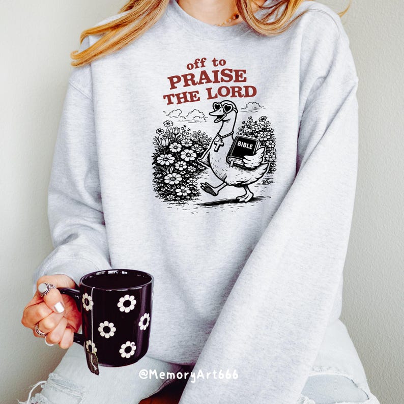 Op de afbeelding: Een lichtgrijs sweatshirt met de tekst "off to PRAISE THE LORD" in bordeauxrood. De afbeelding toont een cartoon gans met een zonnebril en een kruis, die een bijbel draagt. De gans loopt door een bloemenveld.