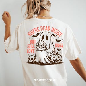 May include: White t-shirt with a graphic of a ghost holding a dog. The text on the shirt reads "You're Dead Inside But Still Love Dogs".
