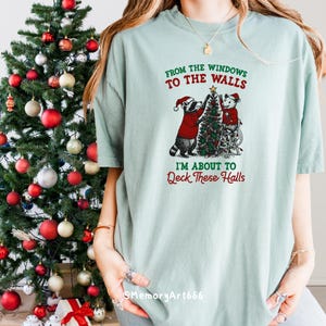 May include: A light green t-shirt with the text "FROM THE WINDOWS TO THE WALLS, I'M ABOUT TO DECK THESE HALLS." The shirt features a graphic of two raccoons decorating a Christmas tree. A decorated Christmas tree is on the left.