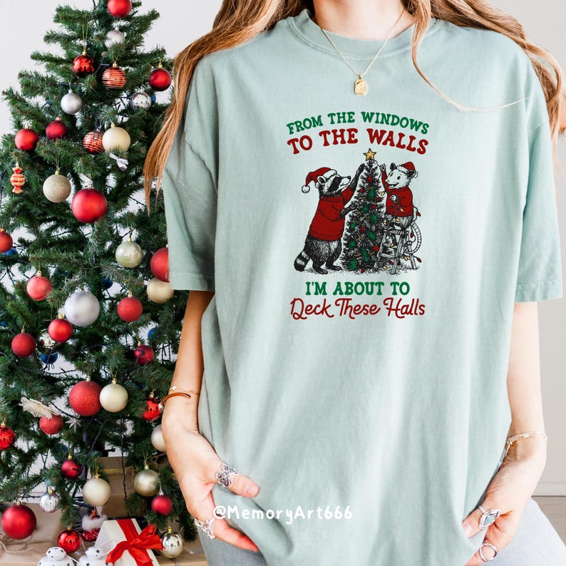 May include: A light green t-shirt with the text "FROM THE WINDOWS TO THE WALLS, I'M ABOUT TO DECK THESE HALLS." The shirt features a graphic of two raccoons decorating a Christmas tree. A decorated Christmas tree is on the left.