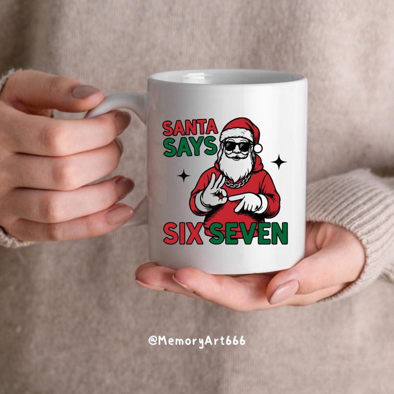 May include: White ceramic mug featuring a graphic of Santa Claus wearing sunglasses and a red hoodie. The text "SANTA SAYS SIX SEVEN" is printed in green and red. The mug is held by a person.