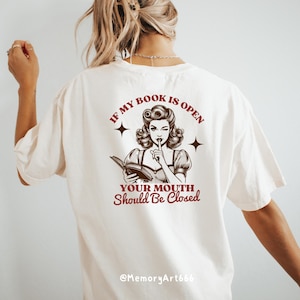 May include: White t-shirt with a brown illustration of a woman reading a book and the text "If my book is open your mouth should be closed" and "@MemoryArt666".