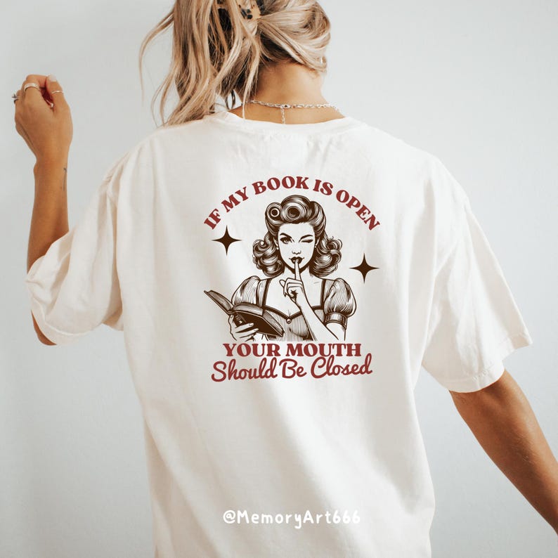 May include: White t-shirt with a brown illustration of a woman reading a book and the text "If my book is open your mouth should be closed" and "@MemoryArt666".