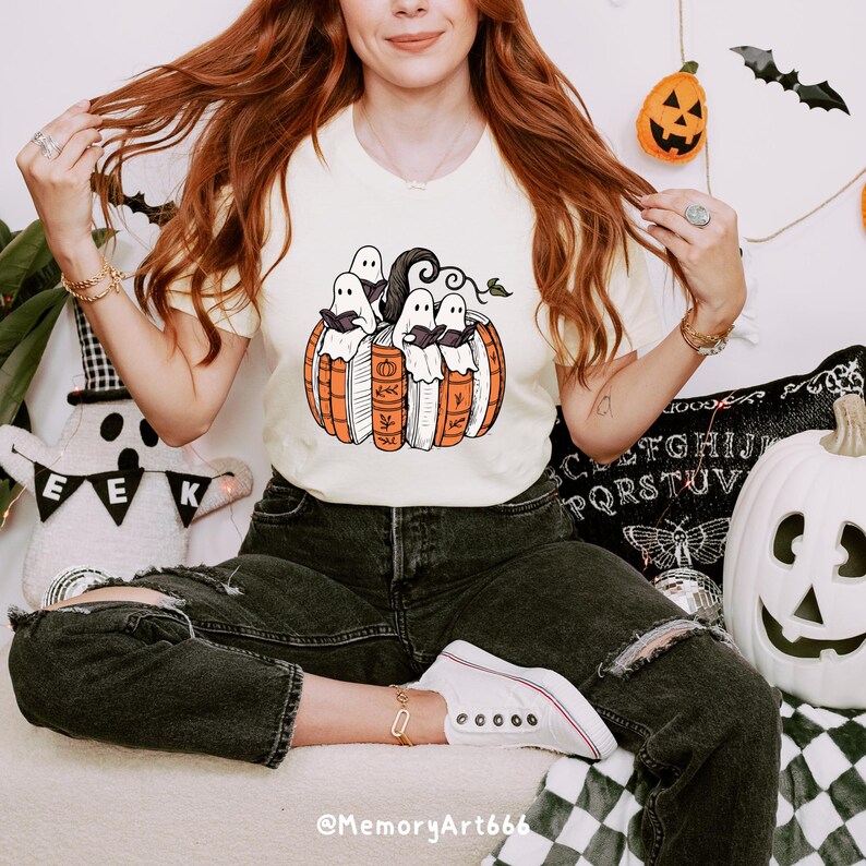 May include: Cream-colored t-shirt featuring a Halloween-themed graphic. The design includes a pumpkin with books and ghosts. The shirt is part of a Halloween collection, perfect for seasonal wear. The text "@MemoryArt666" is at the bottom of the image.