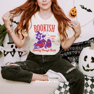 May include: Cream-colored t-shirt with a Halloween-themed graphic. The graphic features a witch reading a book, a cauldron, and the text "BOOKISH" and "Burning Through Books". The shirt is paired with black jeans and white sneakers.