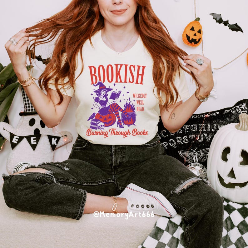 May include: Cream-colored t-shirt with a Halloween-themed graphic. The graphic features a witch reading a book, a cauldron, and the text "BOOKISH" and "Burning Through Books". The shirt is paired with black jeans and white sneakers.