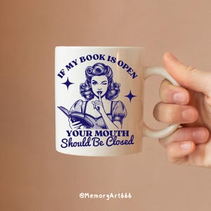 May include: White ceramic mug with a blue illustration of a woman holding a book and putting a finger to her lips. The text on the mug reads "If my book is open your mouth should be closed".