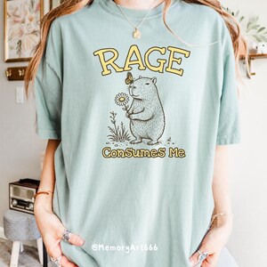 Funny capybara meme PNG SVG design featuring a cute vintage-style capybara holding a daisy flower with a butterfly resting on its head and bold retro “RAGE Consumes Me” typography. Funny sarcastic animal graphic with whimsical hand-drawn line art, soft neutral tones, and trendy unhinged aesthetic. Perfect for funny t-shirts, hoodies, mugs, tote bags, stickers, sublimation projects, Cricut SVG cut files, retro boutique apparel, instant digital download, 300 DPI transparent background.