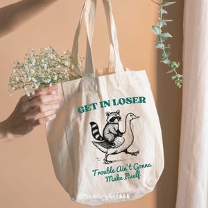 May include: A white canvas tote bag with a black and white illustration of a raccoon riding a goose. The text "GET IN LOSER" is printed in teal above the illustration and "Trouble Ain't Gonna Make Itself" is printed in teal below the illustration.