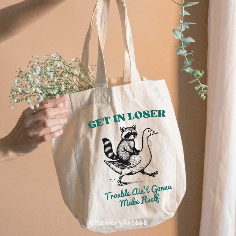 May include: A white canvas tote bag with a black and white illustration of a raccoon riding a goose. The text "GET IN LOSER" is printed in teal above the illustration and "Trouble Ain't Gonna Make Itself" is printed in teal below the illustration.