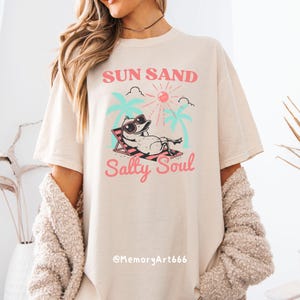 Peut inclure: Un t-shirt beige clair avec un imprim&eacute; graphique d'un canard de dessin anim&eacute; portant des lunettes de soleil et se relaxant sur une chaise de plage. Le texte "Sun Sand Salty Soul" est imprim&eacute; en rose et bleu sur le t-shirt.