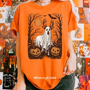 May include: Orange t-shirt featuring a Halloween-themed graphic. The design includes a ghost with deer antlers, a full moon, a bat, and two carved pumpkins. The text "@MemoryArt666" is at the bottom.