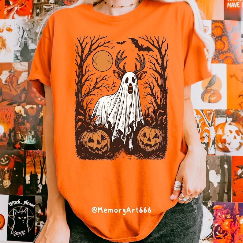 May include: Orange t-shirt featuring a Halloween-themed graphic. The design includes a ghost with deer antlers, a full moon, a bat, and two carved pumpkins. The text "@MemoryArt666" is at the bottom.