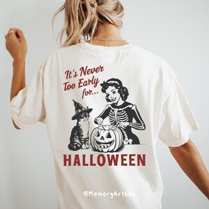Puede incluir: Una camiseta blanca con una ilustraci&oacute;n en blanco y negro de una mujer con un disfraz de esqueleto tallando una calabaza con un gato con un sombrero de bruja a su lado. El texto "It's Never Too Early for... HALLOWEEN" est&aacute; impreso en la camiseta. El texto "@MemoryArt666" est&aacute; impreso en la parte inferior de la camiseta.