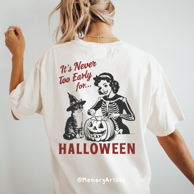 Puede incluir: Una camiseta blanca con una ilustraci&oacute;n en blanco y negro de una mujer con un disfraz de esqueleto tallando una calabaza con un gato con un sombrero de bruja a su lado. El texto "It's Never Too Early for... HALLOWEEN" est&aacute; impreso en la camiseta. El texto "@MemoryArt666" est&aacute; impreso en la parte inferior de la camiseta.