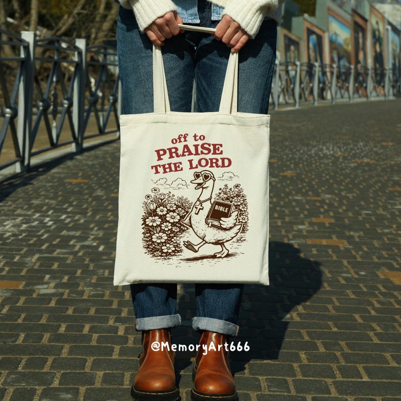 Op de afbeelding: Cr&egrave;mekleurige draagtas met de tekst "off to PRAISE THE LORD" in bordeauxrood. Een cartoon gans met zonnebril en een bijbel is eronder afgedrukt. De gans loopt door een bloemenveld.