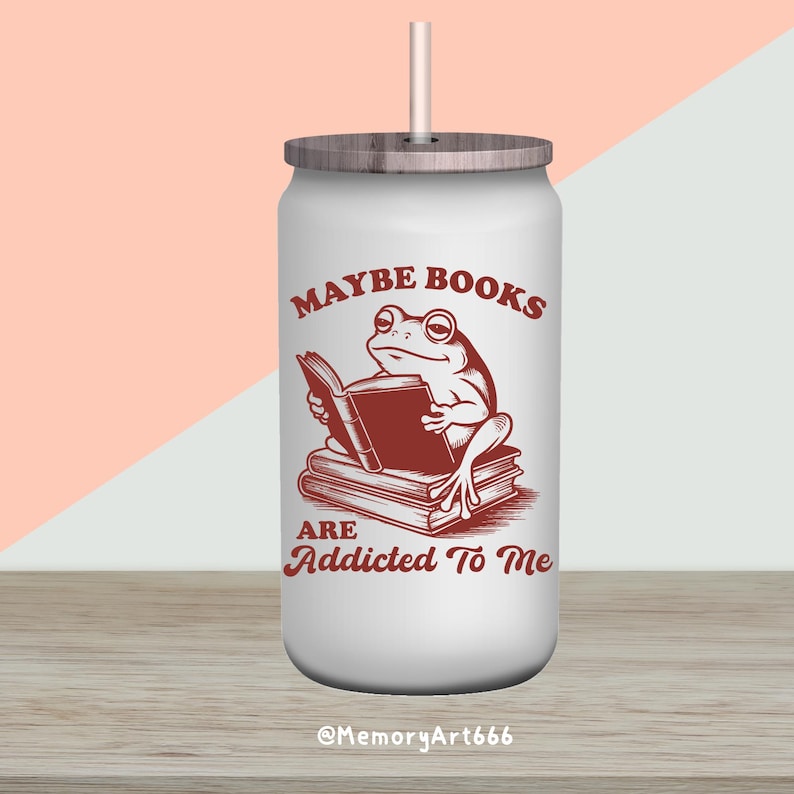 May include: White tumbler with a wooden lid and straw. The tumbler features a brown illustration of a frog reading a book, with the text "Maybe Books Are Addicted To Me". The bottom of the tumbler has the text "@MemoryArt666".