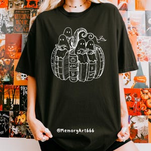 May include: Black t-shirt featuring a white graphic of ghosts reading books inside a pumpkin. The design includes the text "The Witching Hour Near" on the left side. The artist's handle, @MemoryArt666, is printed at the bottom.