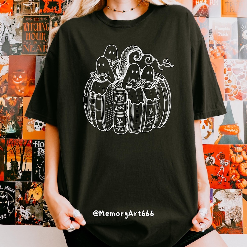 May include: Black t-shirt featuring a white graphic of ghosts reading books inside a pumpkin. The design includes the text "The Witching Hour Near" on the left side. The artist's handle, @MemoryArt666, is printed at the bottom.