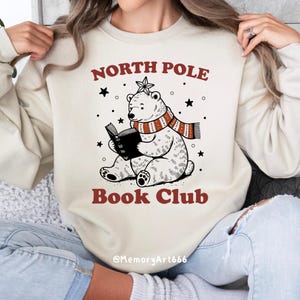May include: A cream-colored sweatshirt with the text "North Pole Book Club" in red and black. A cartoon polar bear wearing a red and white striped scarf is reading a book. The text "@MemoryArt666" is at the bottom of the sweatshirt.