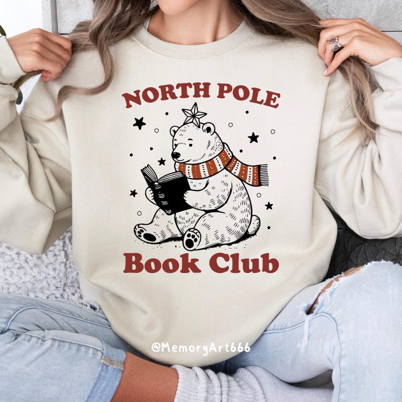 May include: A cream-colored sweatshirt with the text "North Pole Book Club" in red and black. A cartoon polar bear wearing a red and white striped scarf is reading a book. The text "@MemoryArt666" is at the bottom of the sweatshirt.