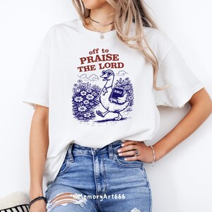 Op de afbeelding: Wit T-shirt met de tekst "off to PRAISE THE LORD" in rood en een blauwe afbeelding van een gans met zonnebril en een bijbel. De gans loopt door een bloemenveld.