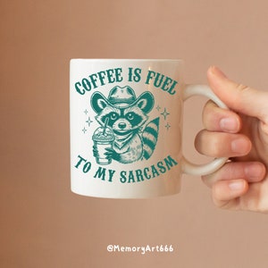 Puede incluir: Taza de cer&aacute;mica blanca con un mapache verde que lleva un sombrero de vaquero y sostiene una taza de caf&eacute;. La taza tiene el texto "Coffee is fuel to my sarcasm" impreso en ella.