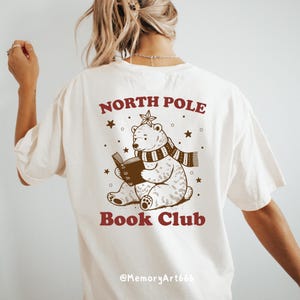 May include: White t-shirt with a brown graphic of a polar bear wearing a scarf and reading a book. The text "North Pole Book Club" is printed on the shirt.