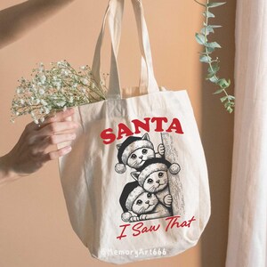 May include: A white canvas tote bag featuring a red and black graphic of three cats wearing Santa hats and peeking around a corner. The text "SANTA I Saw That" is printed in red on the bag.