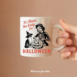 Puede incluir: Taza de cer&aacute;mica blanca con una ilustraci&oacute;n en blanco y negro de una mujer con un disfraz de esqueleto y un gato con un sombrero de bruja. La mujer est&aacute; tallando una calabaza de Halloween. El texto en la taza dice "It's Never Too Early for... HALLOWEEN" y "@MemoryArt666".