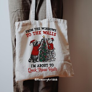 May include: Cream-colored tote bag with a Christmas-themed graphic. The design features a raccoon and a mouse decorating a Christmas tree, with text that reads "From the windows to the walls, I'm about to deck these halls."