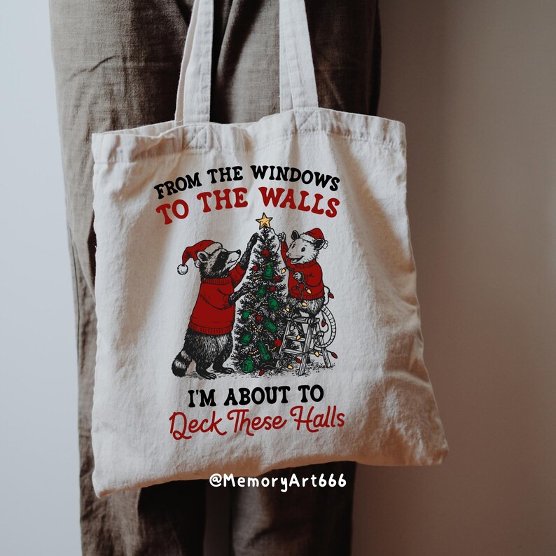 May include: Cream-colored tote bag with a Christmas-themed graphic. The design features a raccoon and a mouse decorating a Christmas tree, with text that reads "From the windows to the walls, I'm about to deck these halls."