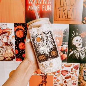 May include: A clear glass tumbler with a bamboo lid and straw, featuring a Halloween-themed design. The design includes a ghost, pumpkins, and a full moon. The tumbler is filled with a light-colored substance. The background includes Halloween-themed posters.