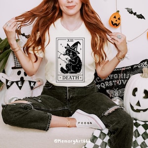 May include: Cream-colored t-shirt featuring a black graphic of a cat wearing a witch hat, with the text "XIII DEATH" in a tarot card design. The cat holds a small object. The shirt is worn with black jeans and white sneakers. Halloween-themed decorations are in the background.