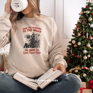 May include: Beige sweatshirt with the text "FROM THE WINDOWS TO THE WALLS I'M ABOUT TO Deck These Halls" and a graphic of two animals decorating a Christmas tree. A decorated Christmas tree is in the background.
