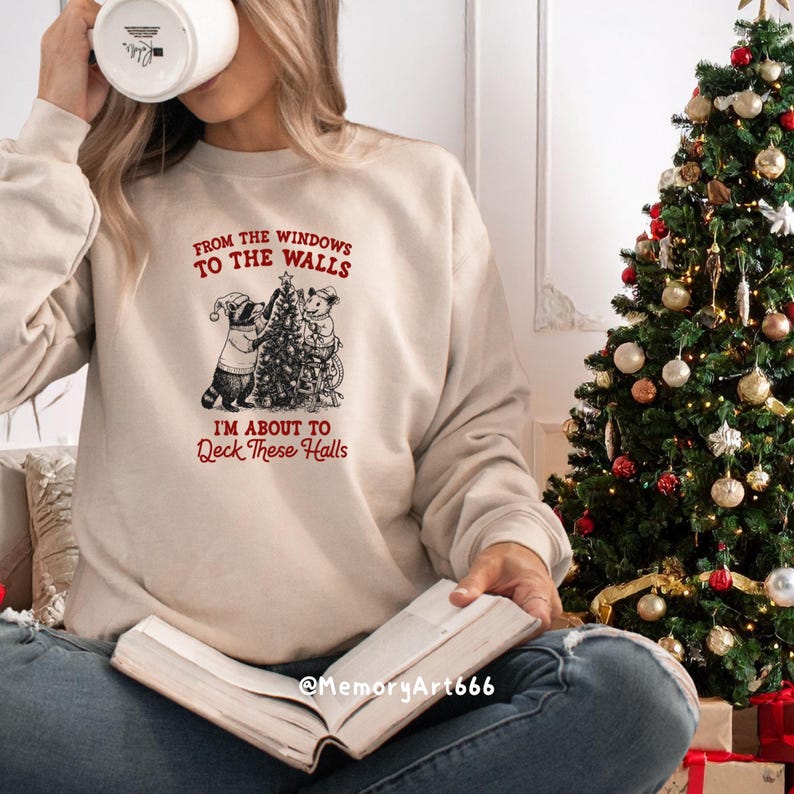 May include: Beige sweatshirt with the text "FROM THE WINDOWS TO THE WALLS I'M ABOUT TO Deck These Halls" and a graphic of two animals decorating a Christmas tree. A decorated Christmas tree is in the background.