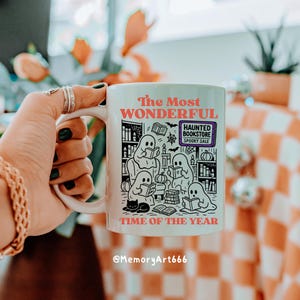 K&ouml;nnte beinhalten: Wei&szlig;e Keramiktasse mit einer umlaufenden Illustration von Geistern, die B&uuml;cher lesen, mit dem Text "The Most Wonderful Time of the Year". Die Tasse zeigt auch den Text "Haunted Bookstore Spooky Sale".