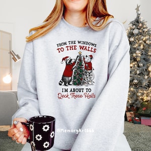 May include: A light gray sweatshirt with a Christmas graphic. The graphic features a raccoon and a mouse decorating a Christmas tree, with the text "FROM THE WINDOWS TO THE WALLS I'M ABOUT TO Deck These Halls". A black mug with white flower designs is held in the hand.