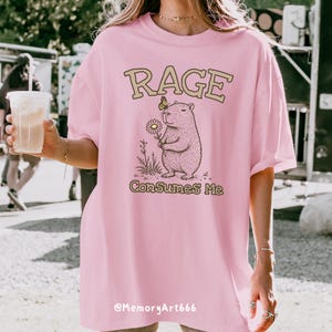Funny capybara meme PNG SVG design featuring a cute vintage-style capybara holding a daisy flower with a butterfly resting on its head and bold retro “RAGE Consumes Me” typography. Funny sarcastic animal graphic with whimsical hand-drawn line art, soft neutral tones, and trendy unhinged aesthetic. Perfect for funny t-shirts, hoodies, mugs, tote bags, stickers, sublimation projects, Cricut SVG cut files, retro boutique apparel, instant digital download, 300 DPI transparent background.