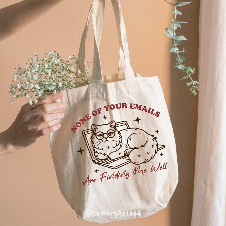 May include: A white canvas tote bag with a brown outline of a cat wearing glasses and lying on a laptop. The text "NONE OF YOUR EMAILS ARE FINDING ME WELL" is printed in red on the bag.