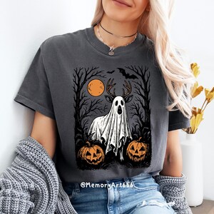 May include: Dark gray t-shirt featuring a Halloween-themed graphic. The design includes a deer-like figure draped in a ghost sheet, set against a backdrop of trees, a full moon, and jack-o'-lanterns. The text "@MemoryArt666" is at the bottom.