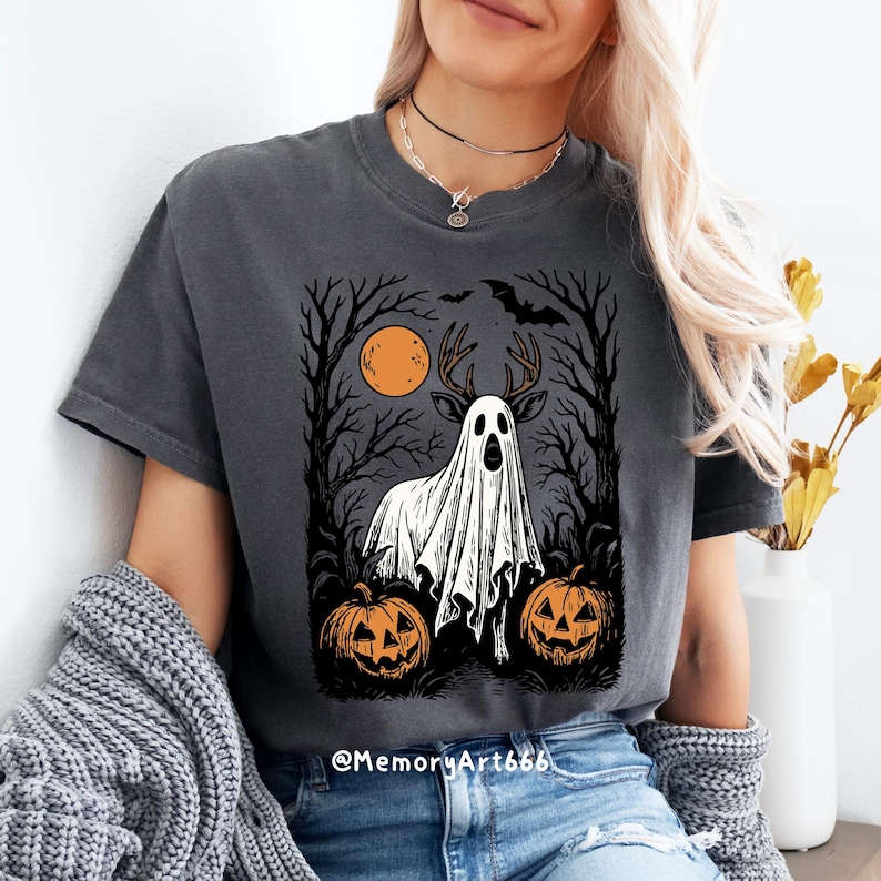 May include: Dark gray t-shirt featuring a Halloween-themed graphic. The design includes a deer-like figure draped in a ghost sheet, set against a backdrop of trees, a full moon, and jack-o'-lanterns. The text "@MemoryArt666" is at the bottom.