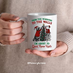 May include: White ceramic mug featuring a festive design with a raccoon and a mouse decorating a Christmas tree. The mug has the text "FROM THE WINDOWS TO THE WALLS, I'M ABOUT TO Deck These Halls."