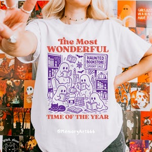 K&ouml;nnte beinhalten: Wei&szlig;es T-Shirt mit einer lila-orangenen Grafik. Die Grafik zeigt Geister, die in einer Buchhandlung lesen, mit dem Text "The Most Wonderful Time of the Year" und "Haunted Bookstore Spooky Sale."