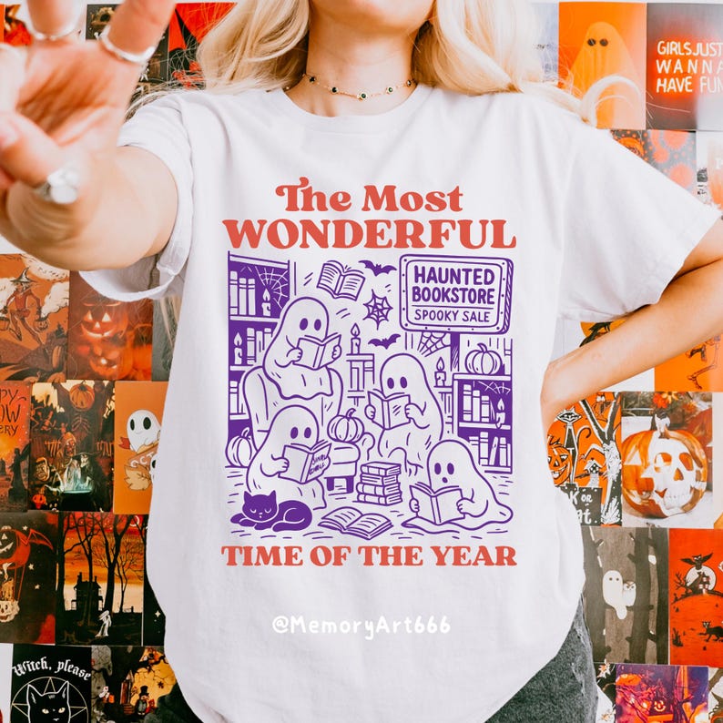 K&ouml;nnte beinhalten: Wei&szlig;es T-Shirt mit einer lila-orangenen Grafik. Die Grafik zeigt Geister, die in einer Buchhandlung lesen, mit dem Text "The Most Wonderful Time of the Year" und "Haunted Bookstore Spooky Sale."