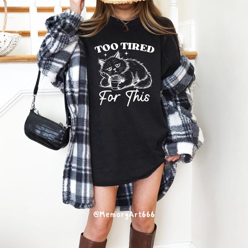 May include: Black t-shirt with the text "TOO TIRED For This" and a white graphic of a cat holding a coffee cup. The person is wearing a black and white plaid jacket and brown boots. A black purse hangs from the shoulder.