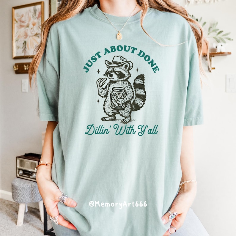 K&ouml;nnte beinhalten: Hellgr&uuml;nes T-Shirt mit einem Cartoon-Waschb&auml;ren, der einen Cowboyhut tr&auml;gt und ein Glas h&auml;lt. Der Text auf dem Shirt lautet "Just About Done" und "Dillin' With Y'all". Das Shirt hat eine lockere Passform.