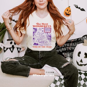K&ouml;nnte beinhalten: Cremefarbenes T-Shirt mit einem Halloween-Motiv. Das Motiv zeigt Geister, die in einer Buchhandlung lesen, mit dem Text "The Most Wonderful Time of the Year". Das Shirt wird mit schwarzen Jeans und wei&szlig;en Turnschuhen kombiniert.