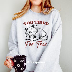 May include: A light gray sweatshirt with the text "TOO TIRED For This" and a graphic of a grumpy cat holding a coffee cup. The cat is drawn in black and white. A black mug with white flower designs is held in the hand.