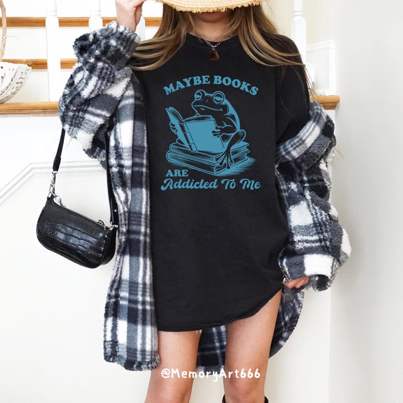 May include: Black long-sleeve shirt with the text "MAYBE BOOKS ARE ADDICTED TO ME" and a blue frog reading a book. The shirt is worn with a black and white plaid jacket and a black purse.